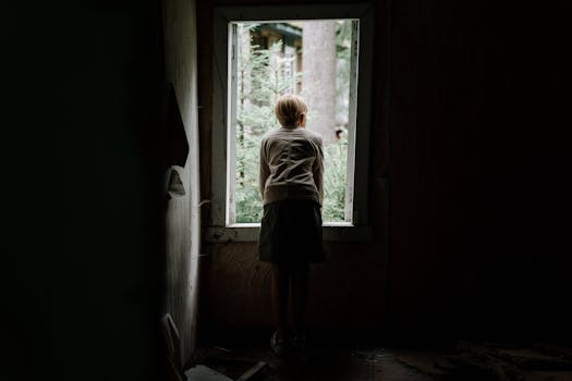 A young child curiously looks through a window of an abandoned building into the greenery outside.